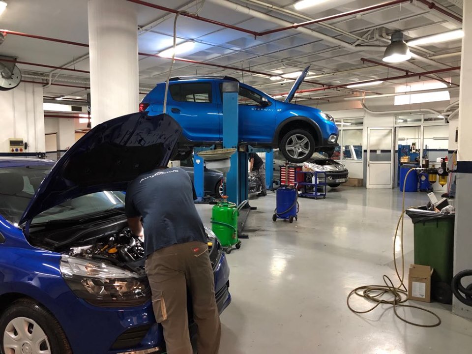 a man wearing a blue t-shirt and brown pants fixing a blue car while behind him there is another car to fix