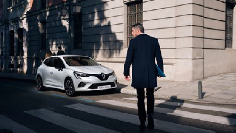 a man with casual look is walking down the street heading to a white reanult clio