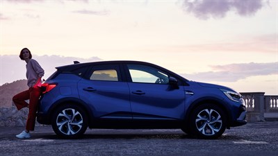 A blue Renault Captur from the side view in a mountain and a woman is standing in the backside of the car