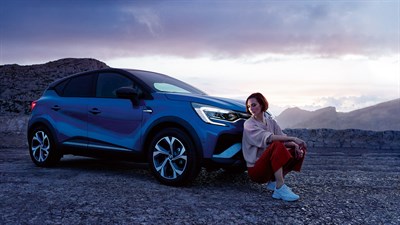 A blue Renault Captur from the side view in a mountain and a woman is standing in the front of the car