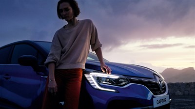 A blue Renault Captur from the side view in a mountain and a woman is standing in the front right side of the car